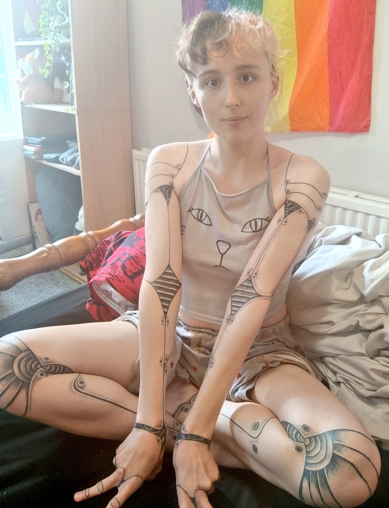 trans girl wearing a gray cami with a cat face graphic, tattooed with external mechanical plating